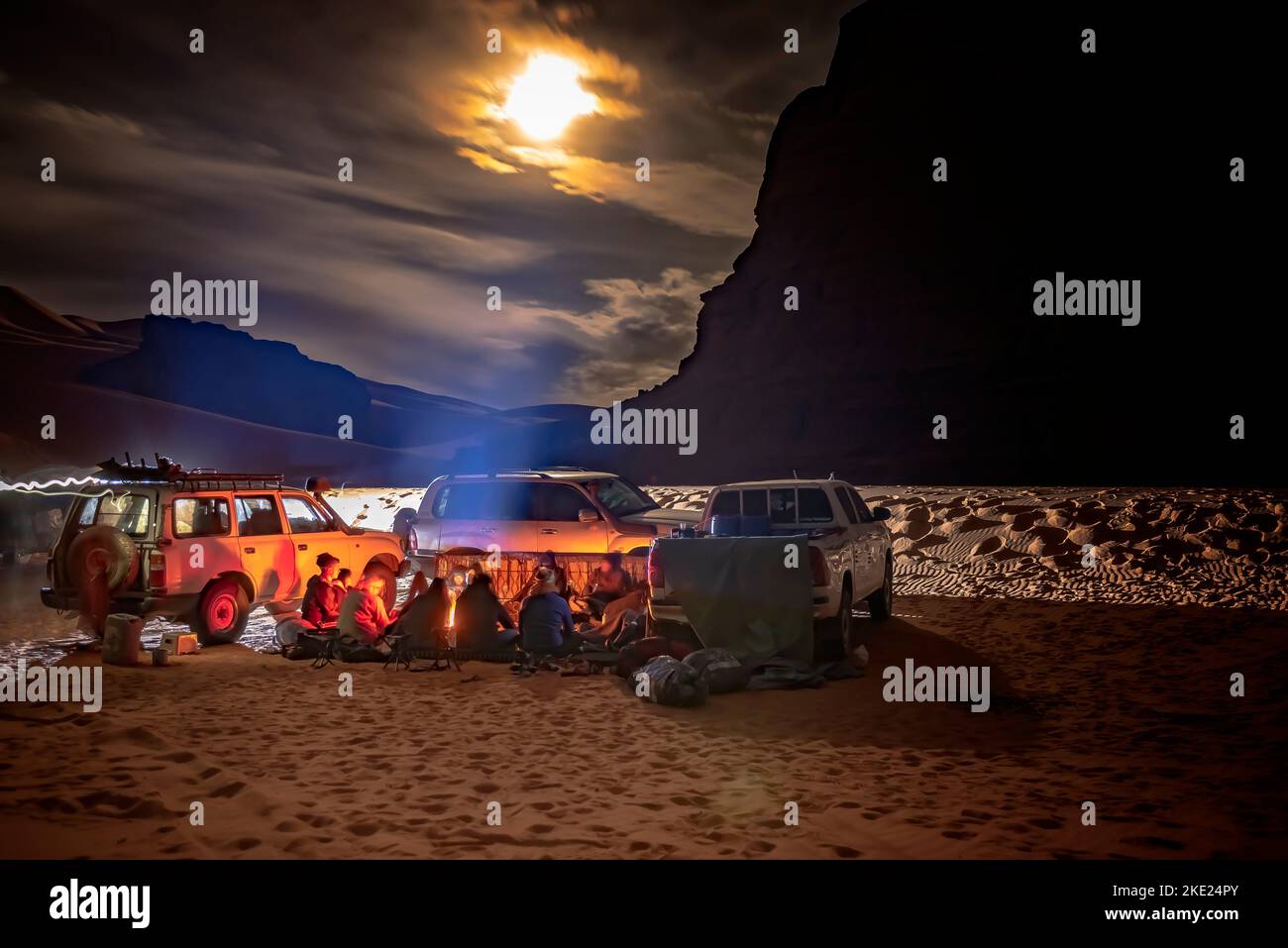 Camping group in Sahara Desert by night. 4X4 vehicules parked, tourists and local berber people sitting around a campfire. cloudy sky moonlight. Stock Photo