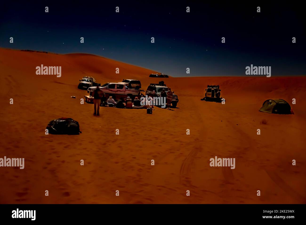 Unrecognizable group of tourists and tuareg camping in Sahara Desert by night. 4X4 vehicles parked, red colored sand dunes, stars in dark sky. Stock Photo