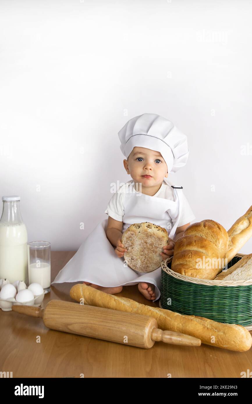 A little cook boy in an apron and a cap sits on a table with a bun in his hands. Stock Photo