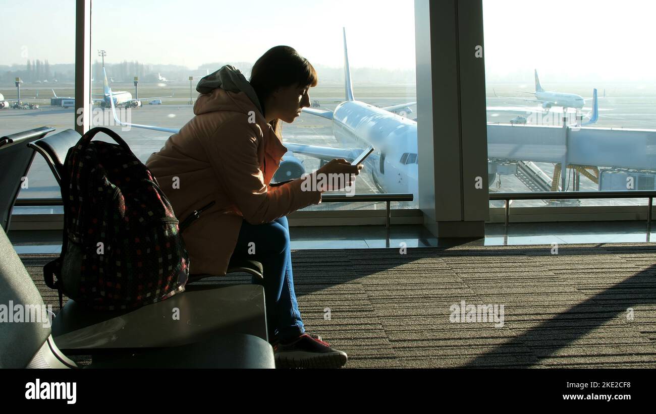 at the airport, in the waiting room, against the background of a window overlooking the airplanes and the runway, sits a young woman typing on the phone. see her silhouette. High quality photo Stock Photo