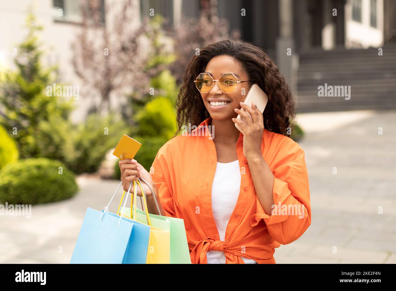 Cheerful millennial black lady in casual, glasses with bags of purchases with credit card, calls by phone Stock Photo