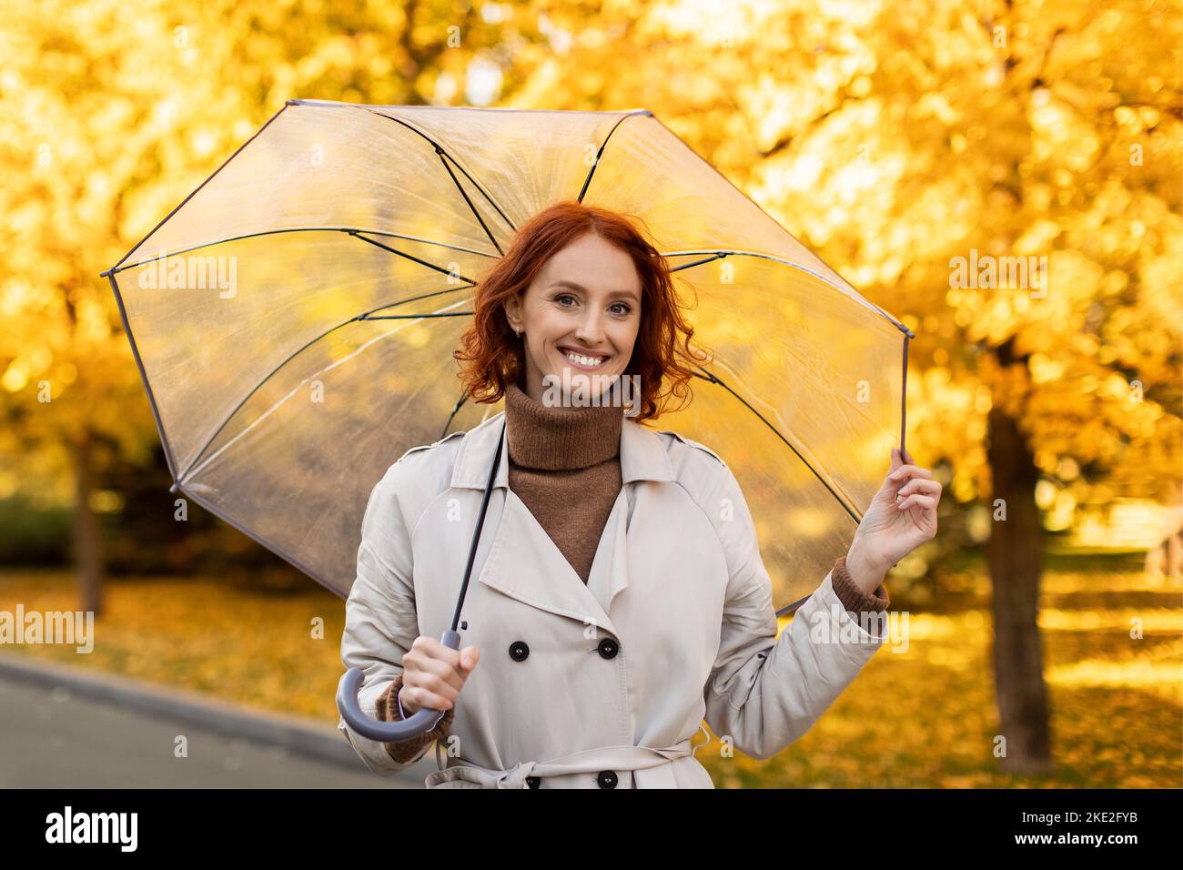 Happy caucasian millennial woman with red hair in raincoat with umbrella enjoys rain, weather and yellow leaves Stock Photo
