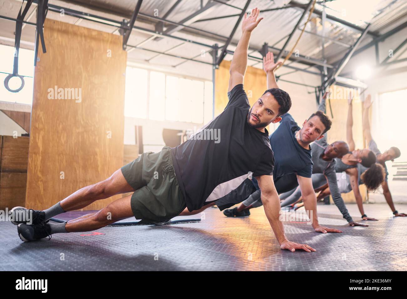 Strong man, personal trainer and group training with side plank exercise, workout and fitness in gym club. Bodybuilder coach teaching sports class Stock Photo