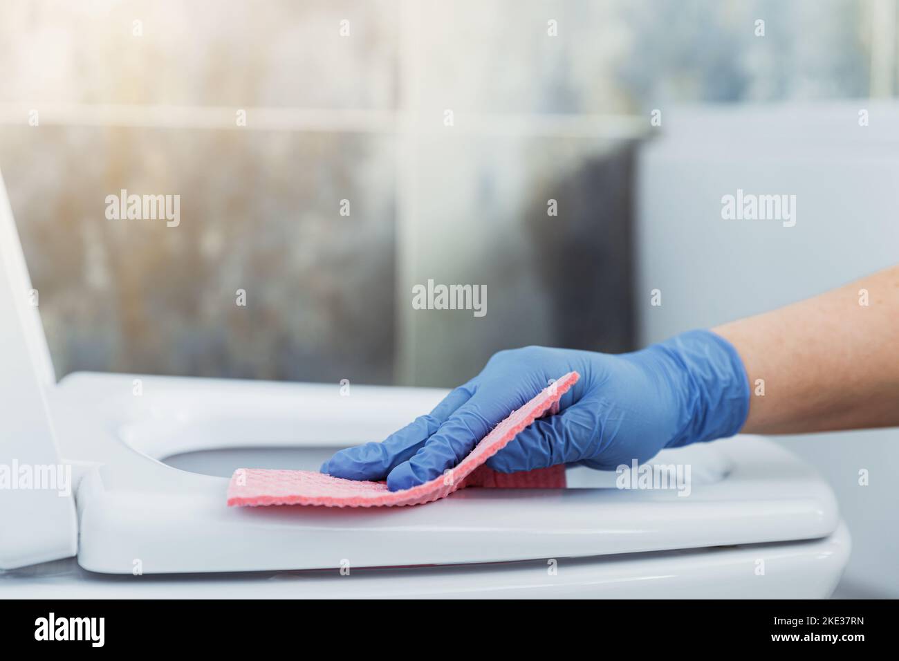 Cropped image view with close up on woman hands gloved in rubber protective gloves. Housewife cleaning toilet bowl, seat with pink cloth, wipe in bathroom or modern public restroom with blue grey tile Stock Photo