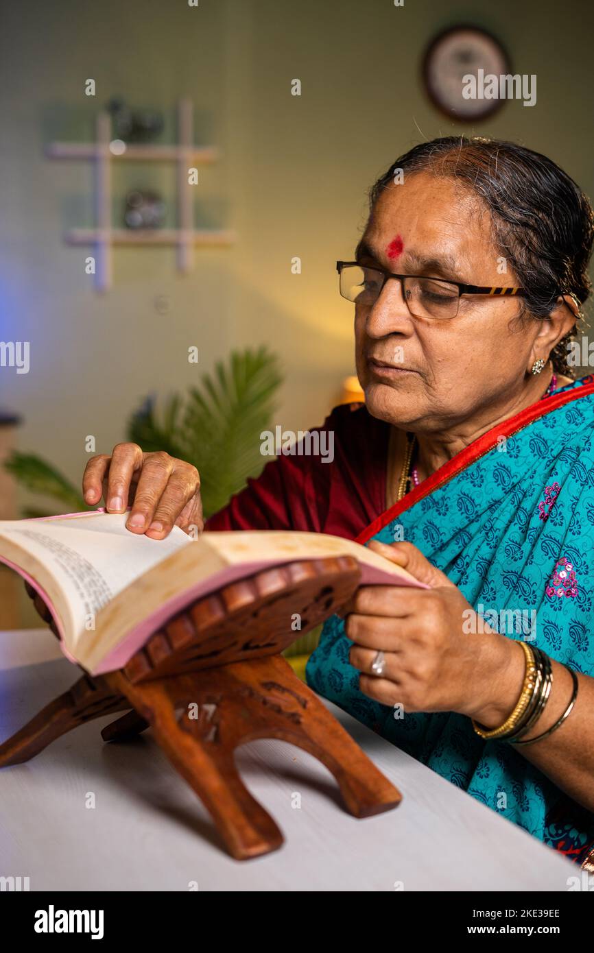 vertical shot of Senior Indian woman with eyeglasses reading novel at home - concept of leisure activity, hobbies and knowledge. Stock Photo