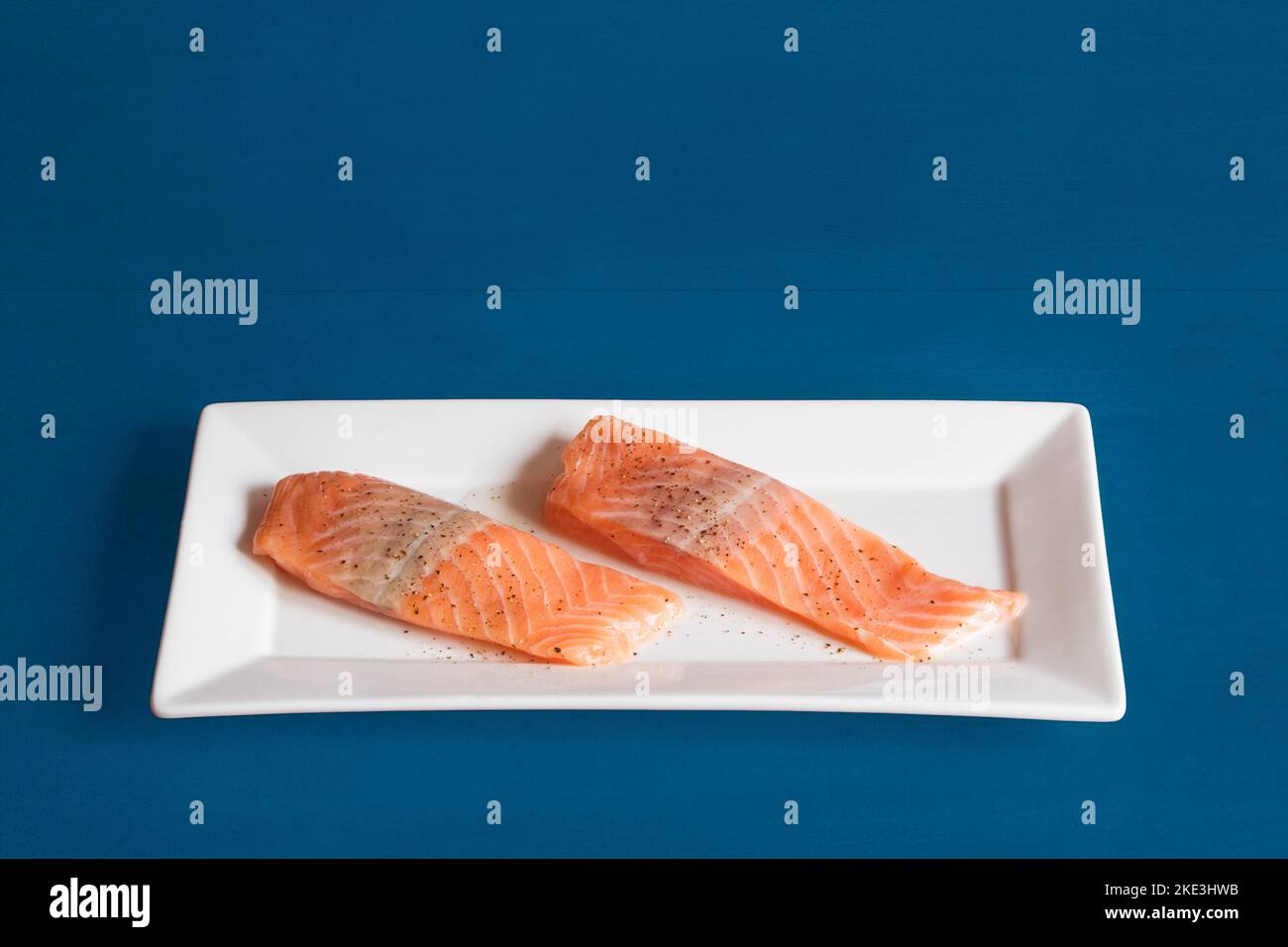 Two raw salmon loins seasoned with salt and pepper on a rectangular white ceramic platter on a blue studio background with copy space. Food and fish. Stock Photo
