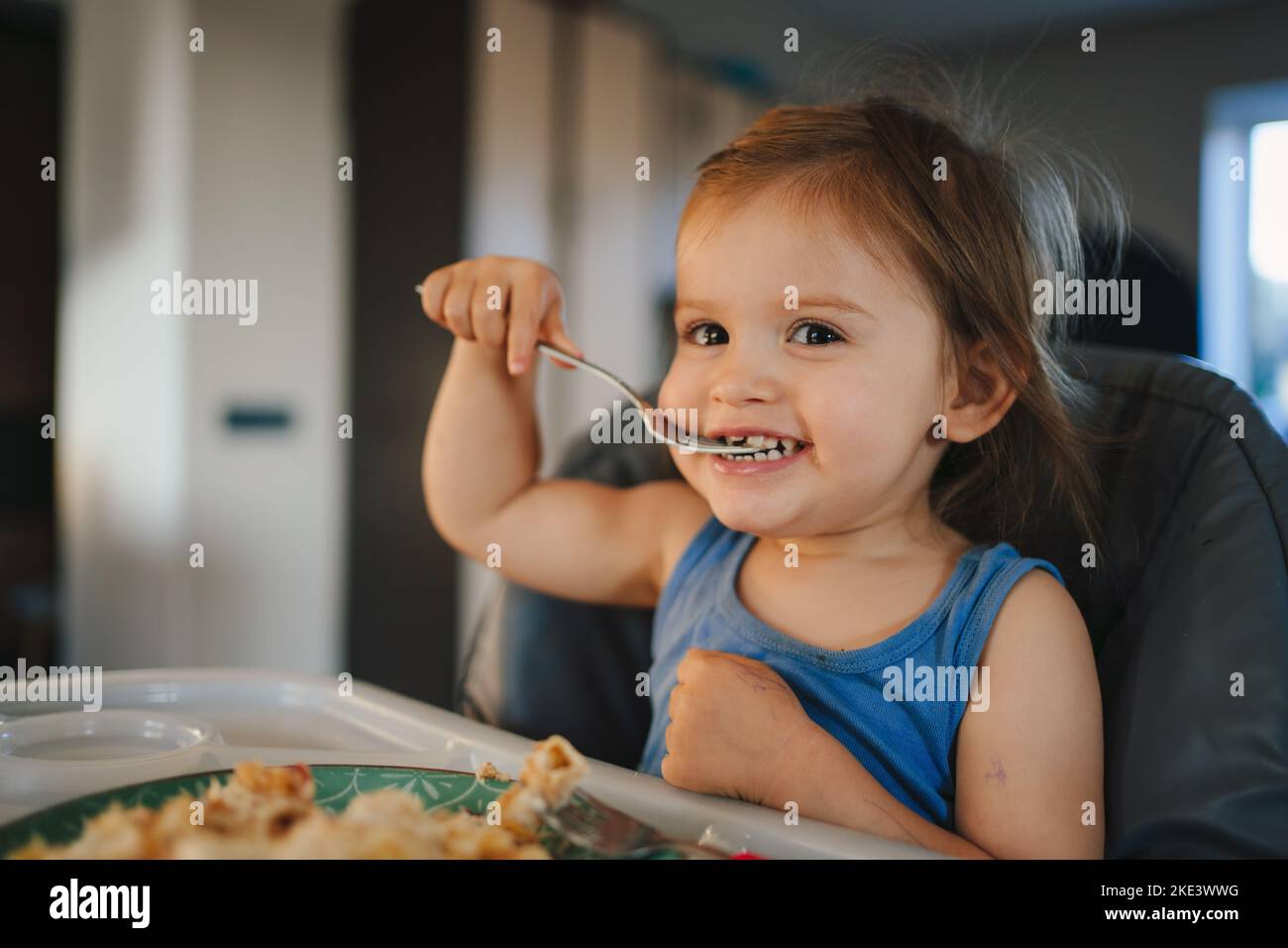 Baby sitting in highchair with spoon and plate on kitchen. Eating food