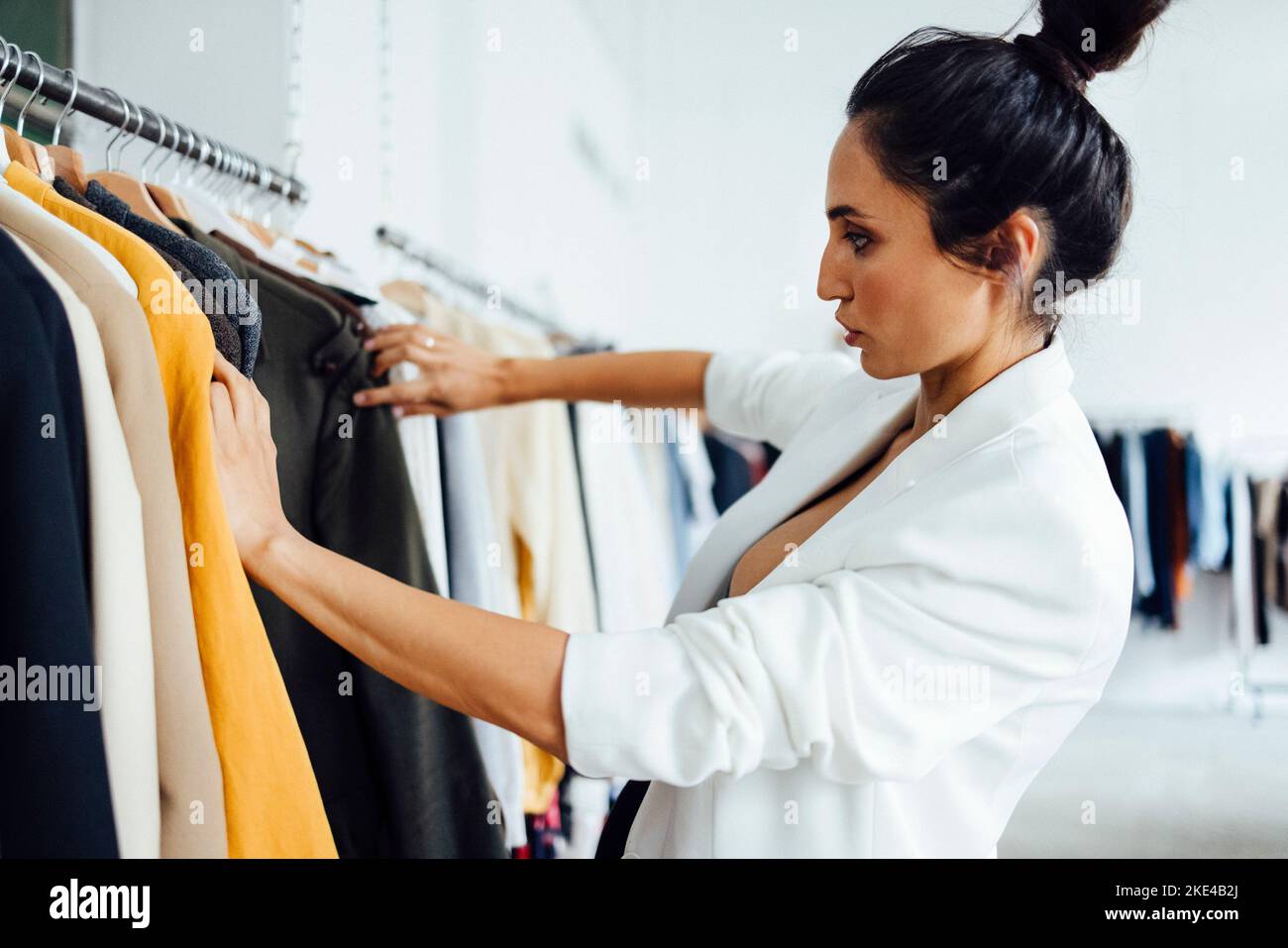 Pretty young woman standing in front of a hanger and trying to choose clothes for work or outing. Selection of wardrobe, stylist, shopping. Stock Photo
