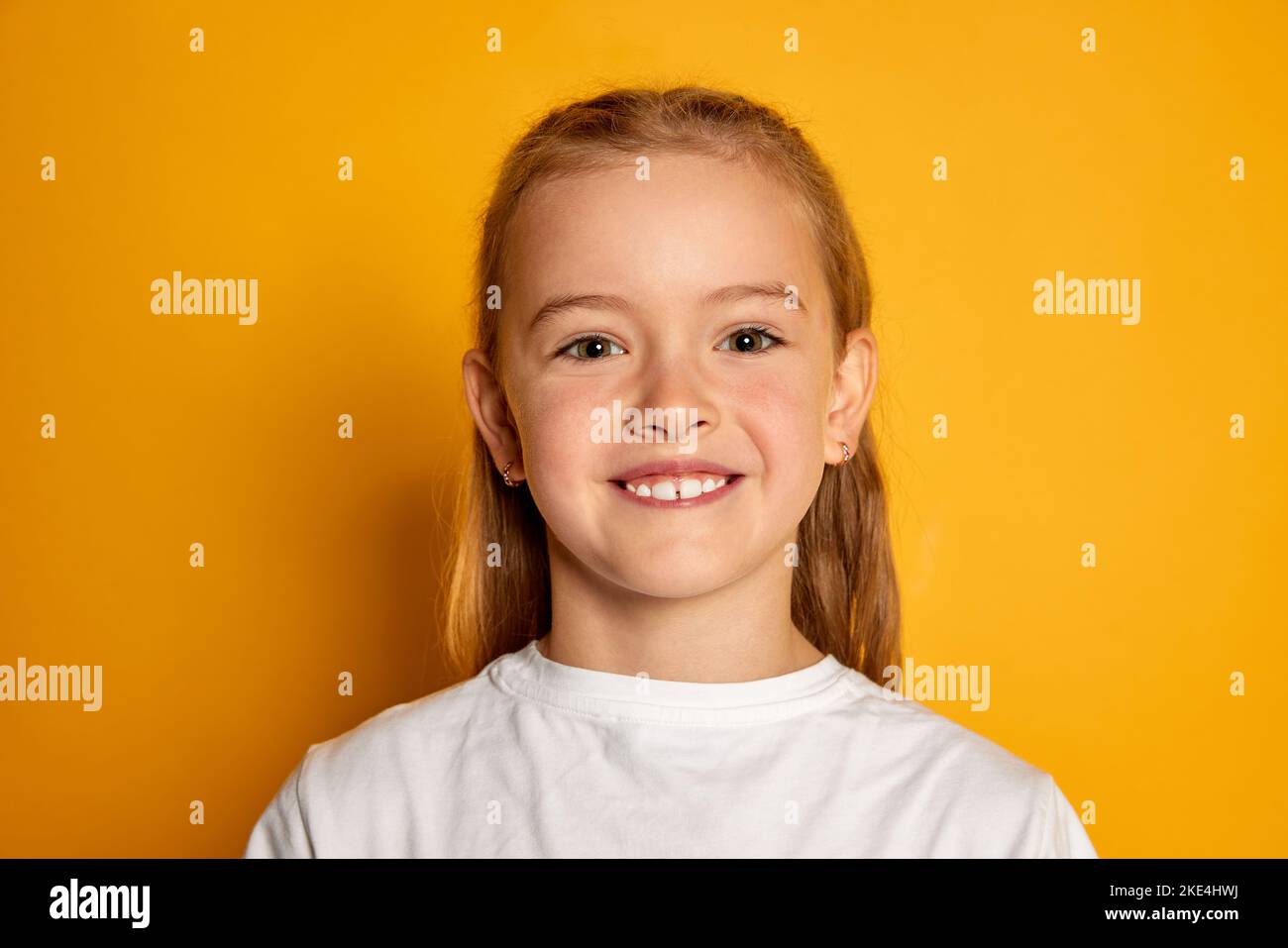 Portrait of little beautiful girl, child in white T-shirt posing, smiling at camera isolated over yellow studio background Stock Photo