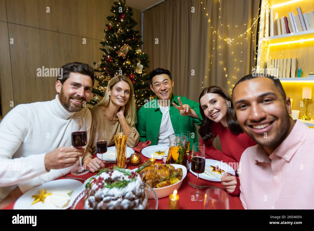 Festive selfie of a group of interracial people. Young men and women are sitting at the table in the apartment near the Christmas tree, holding glasses, looking at the camera, smiling. Stock Photo