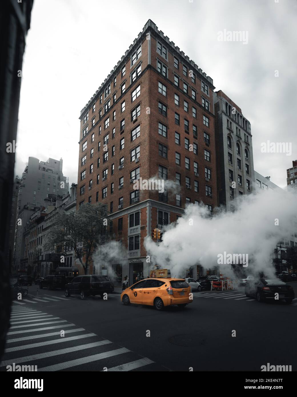 A high rise building in midtown manhattan and a yellow cab passing in on the intersection in front of it with steam coming out of a manhole cover Stock Photo