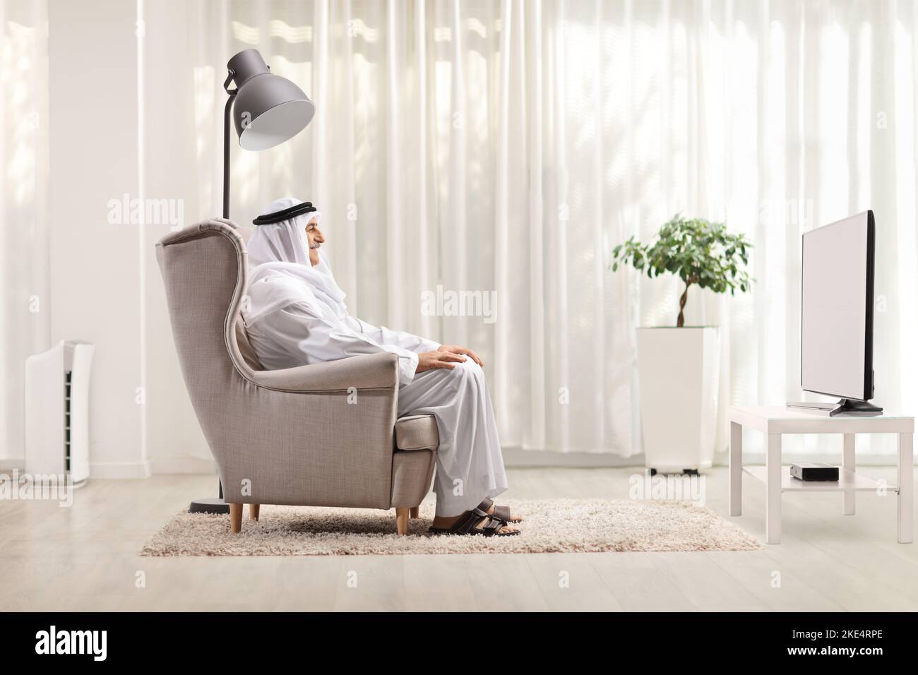 Arab man in ethnic clothes sitting in an armchair and watching tv at home Stock Photo