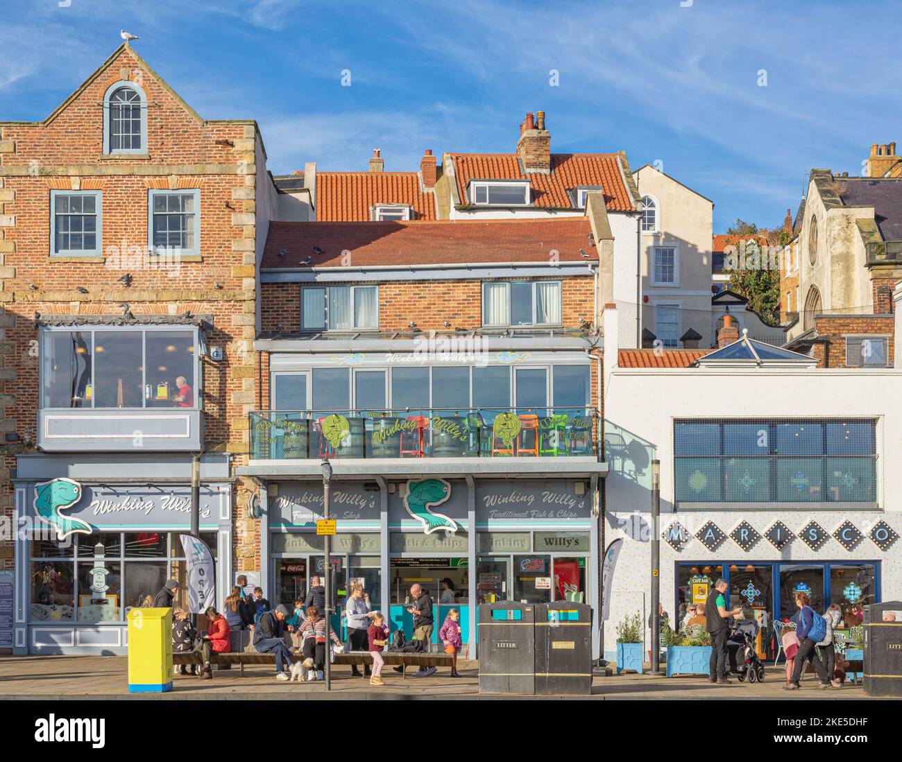A typical view of a fish and chip restaurant at the seaside. People sit on a bench eating and a blue sky is above. Stock Photo