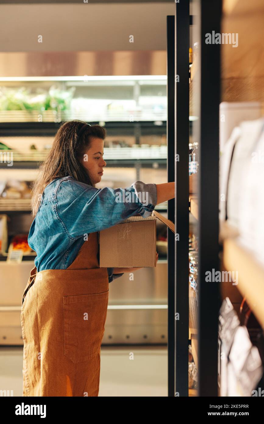 Grocery store worker with Down syndrome restocking food products in a shop. Empowered woman with an intellectual disability working as a shopkeeper in Stock Photo