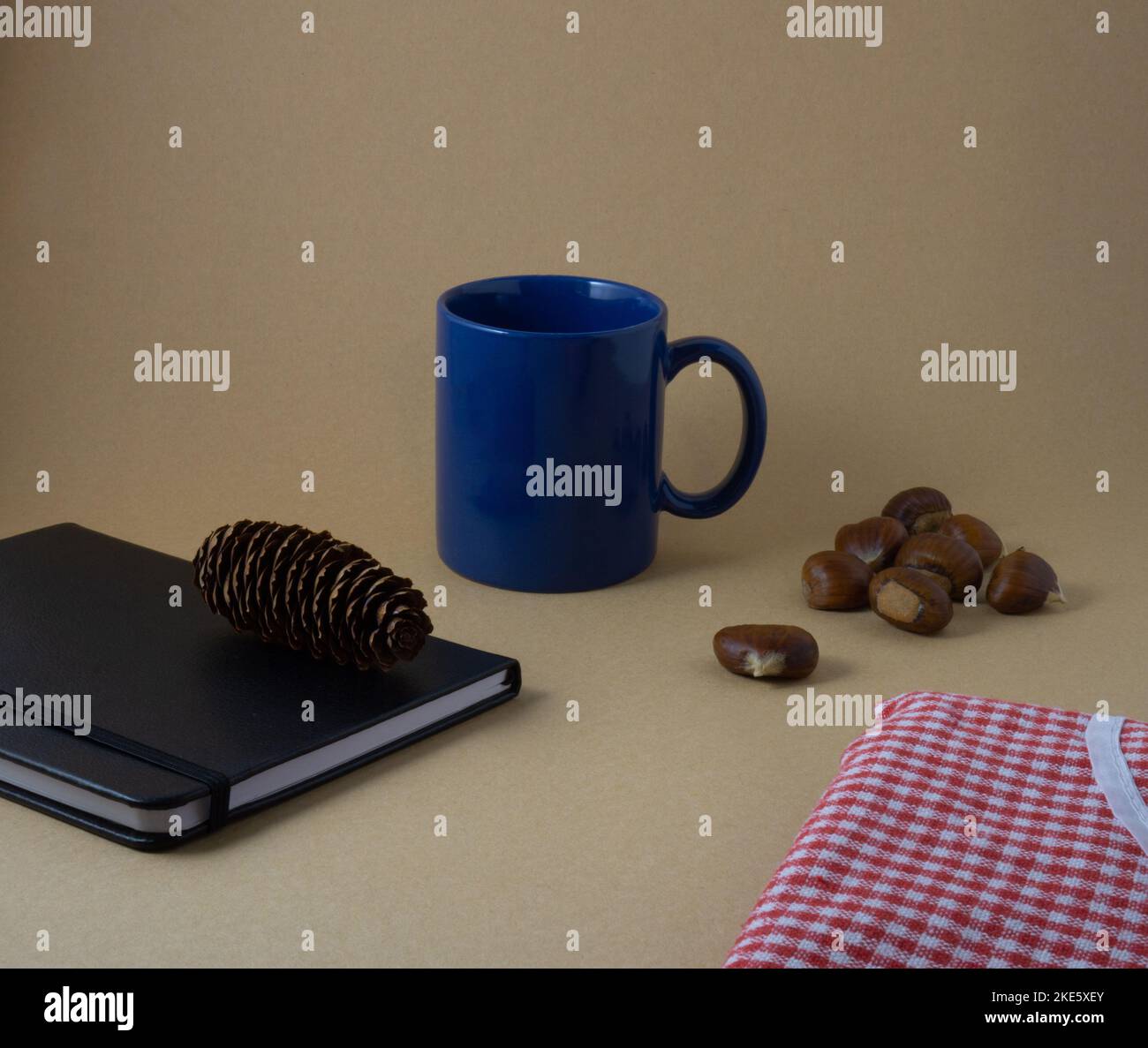 autumn composition with blue cup, black notebook with pine cone on top, chestnuts and red blanket Stock Photo