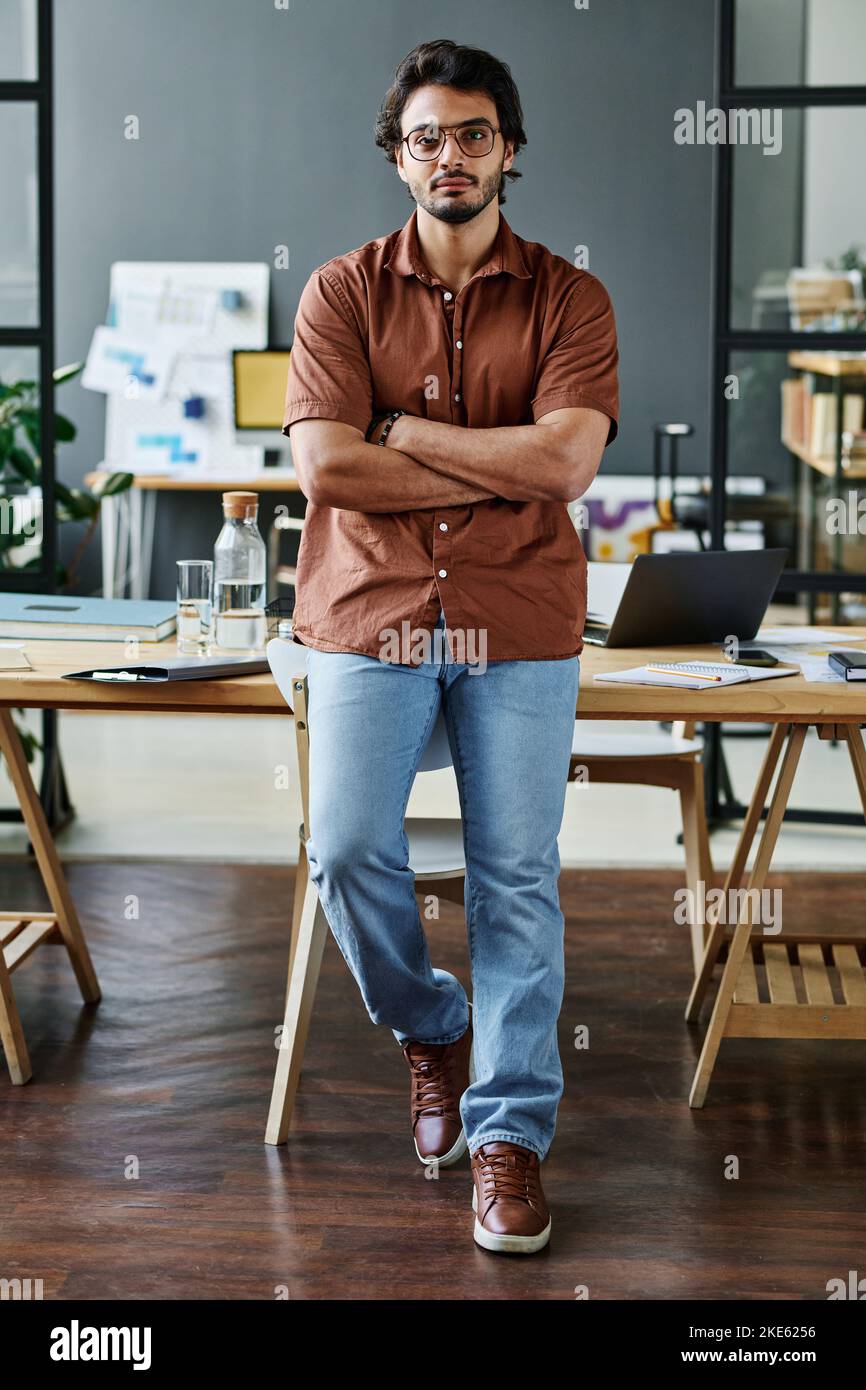Young confident businessman in smart casualwear and eyeglasses crossing arms on chest while standing against his workplace Stock Photo
