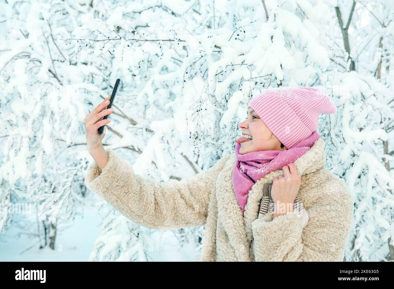 A young woman on the background of a beautiful plant in the snow. A young lady shows her tongue to the phone. Great mood, joy, fun. Stock Photo