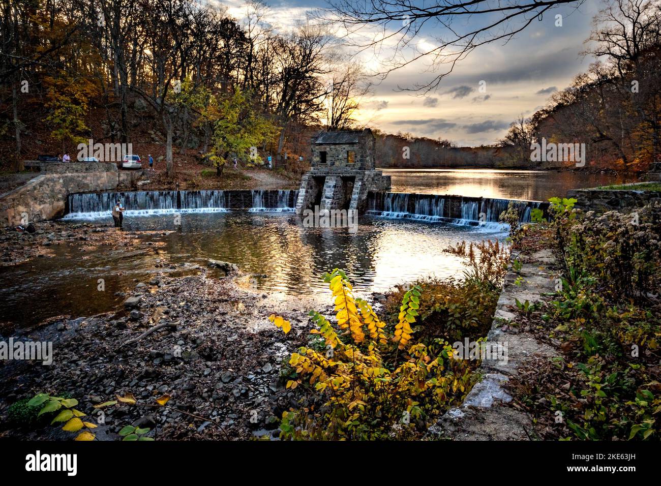 Morristown, NJ - USA - Nov 5, 2022 An autumnal horizontal wide angle view of New Jersey's historic stone Speedwell Dam during sunset. Fly fisherman in Stock Photo