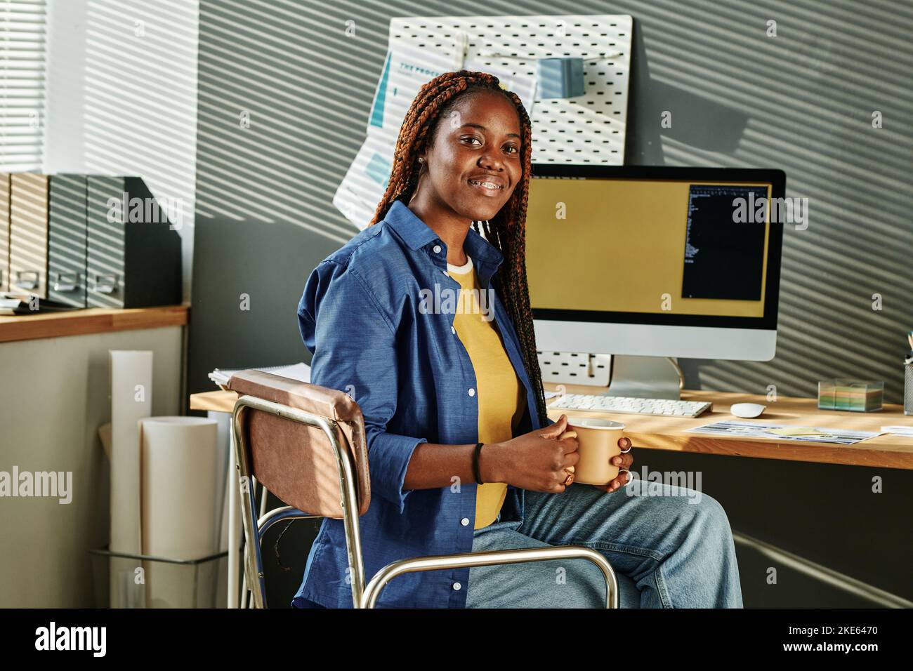 Happy young African American female intern with cup of coffee looking at camera while sitting by workplace with computer monitor Stock Photo