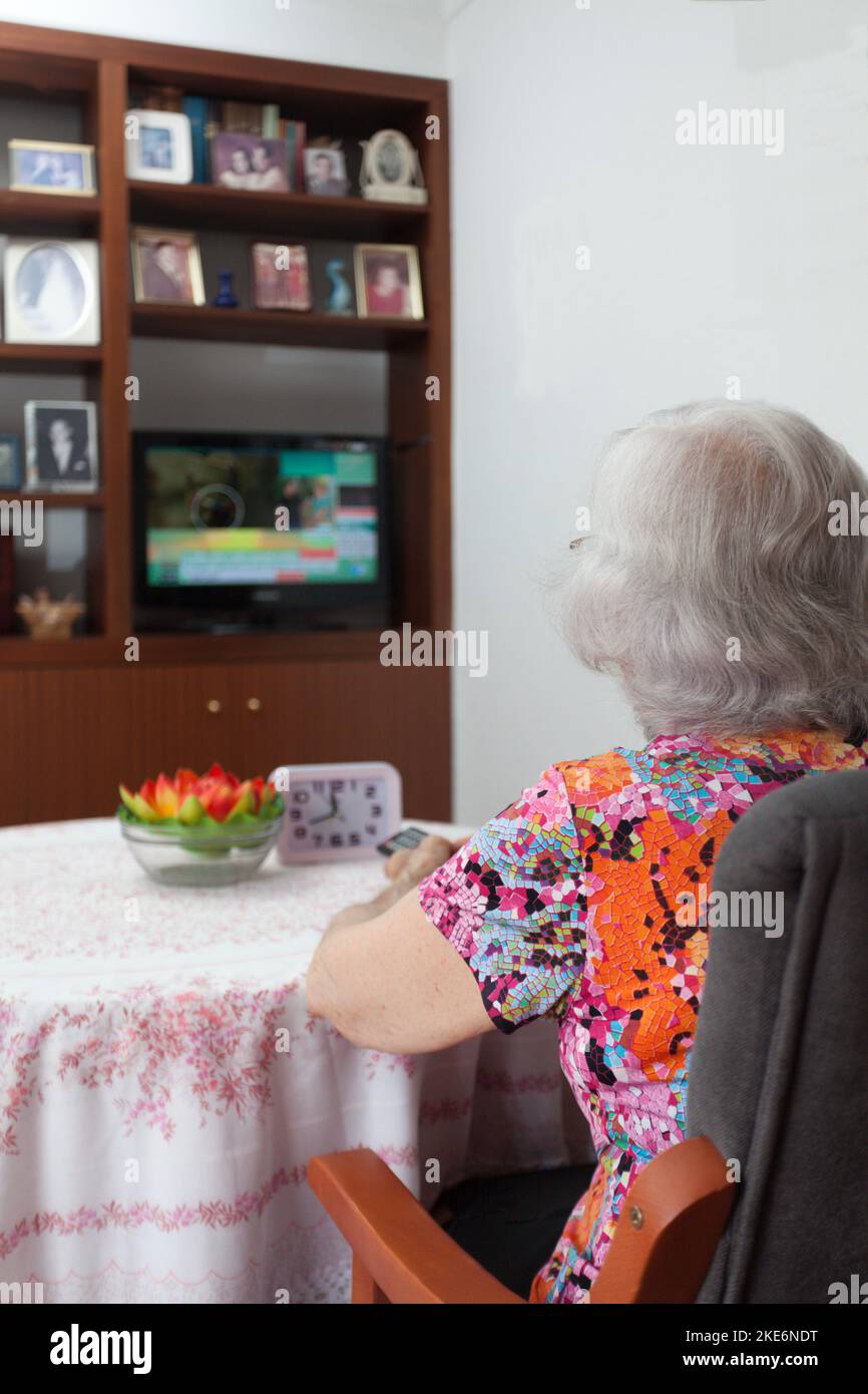 older white-haired woman watching TV in her living room, vertical,  loneliness of the elderly, connectivity with the outside world. Stock Photo