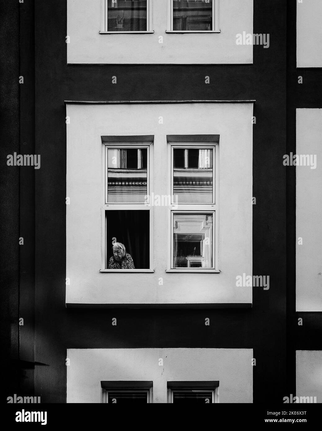 A vertical grayscale shot of an old lady looking out of the window. Vienna, Austria Stock Photo