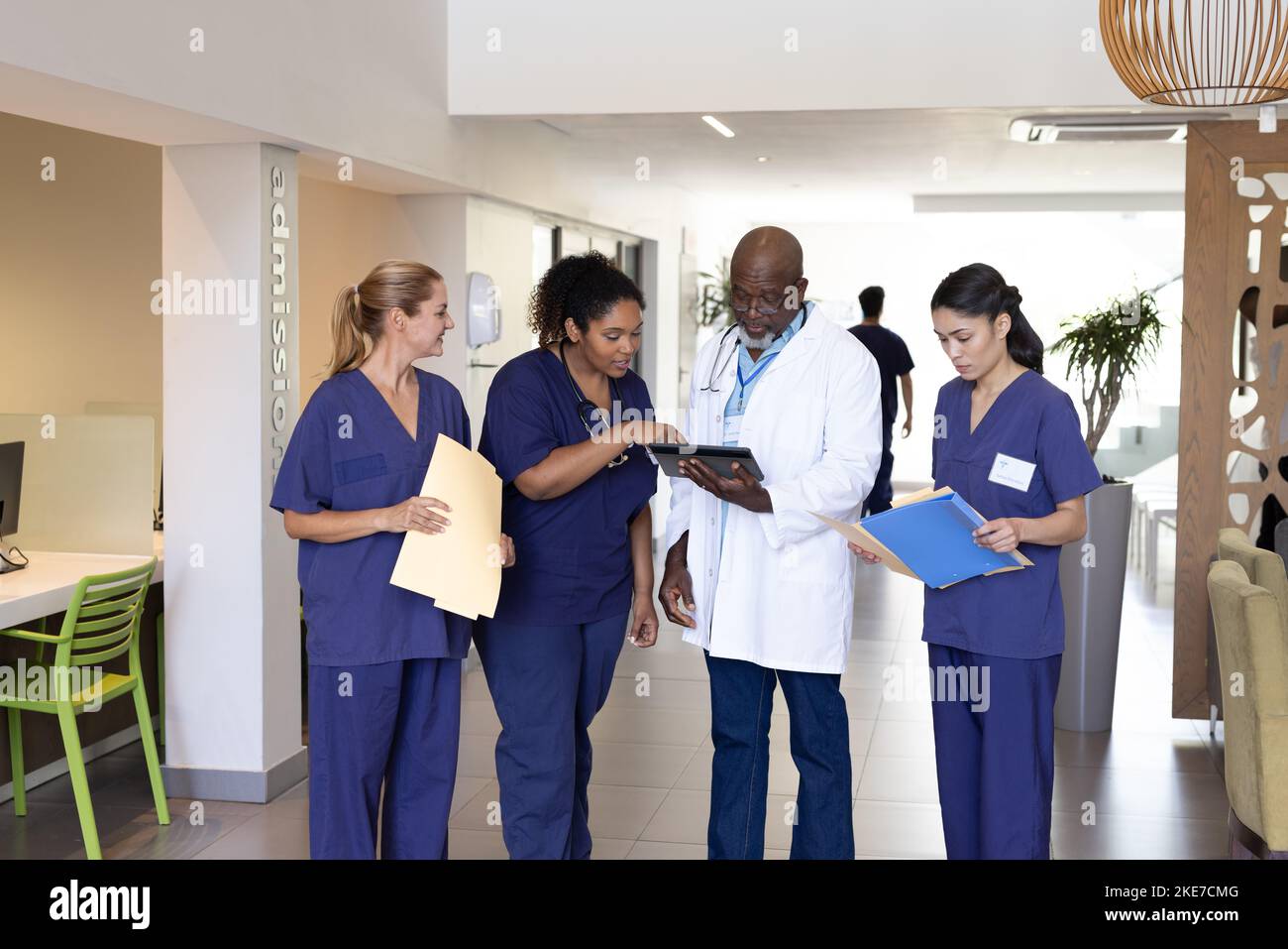 Diverse group of healthcare workers with tablet and paperwork discussing in hospital corridor Stock Photo