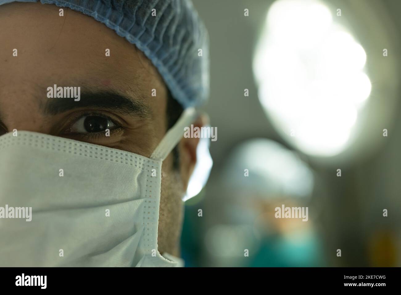 Portrait of biracial male surgeon in surgical cap and mask in operating ...