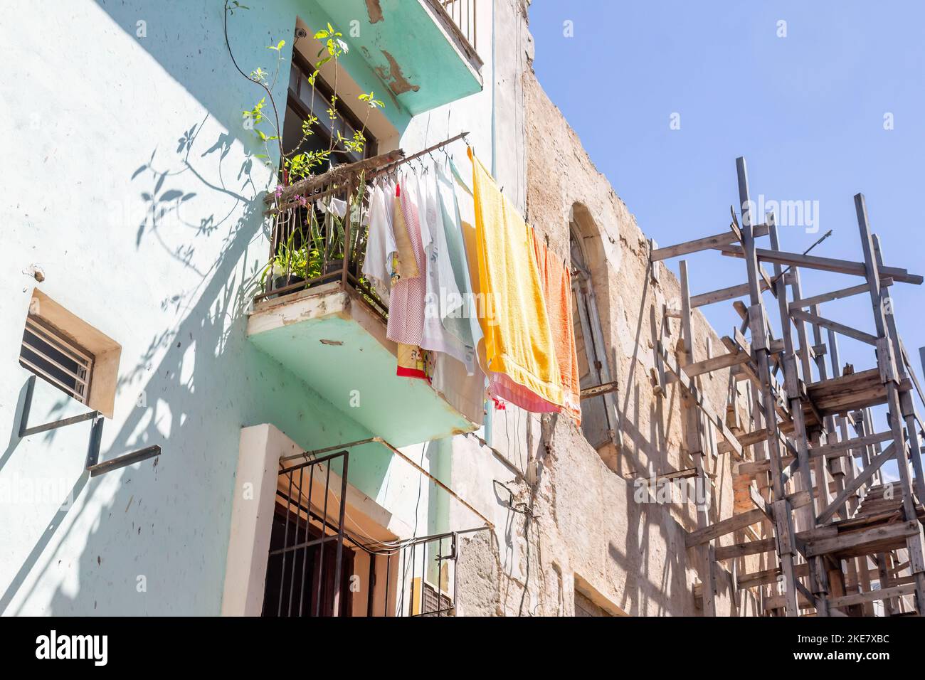 Clothes are drying in the wind in the balcony of an apartment building. On the side, there is a collapsing building with support wood scaffolding. Stock Photo