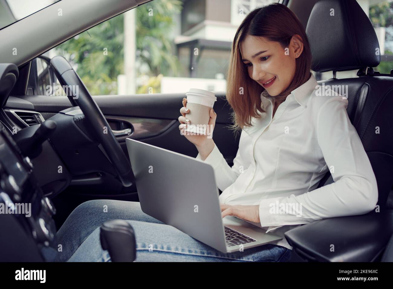Young woman using laptop and drinks coffee at the wheel of the car. Multitasking. Stock Photo