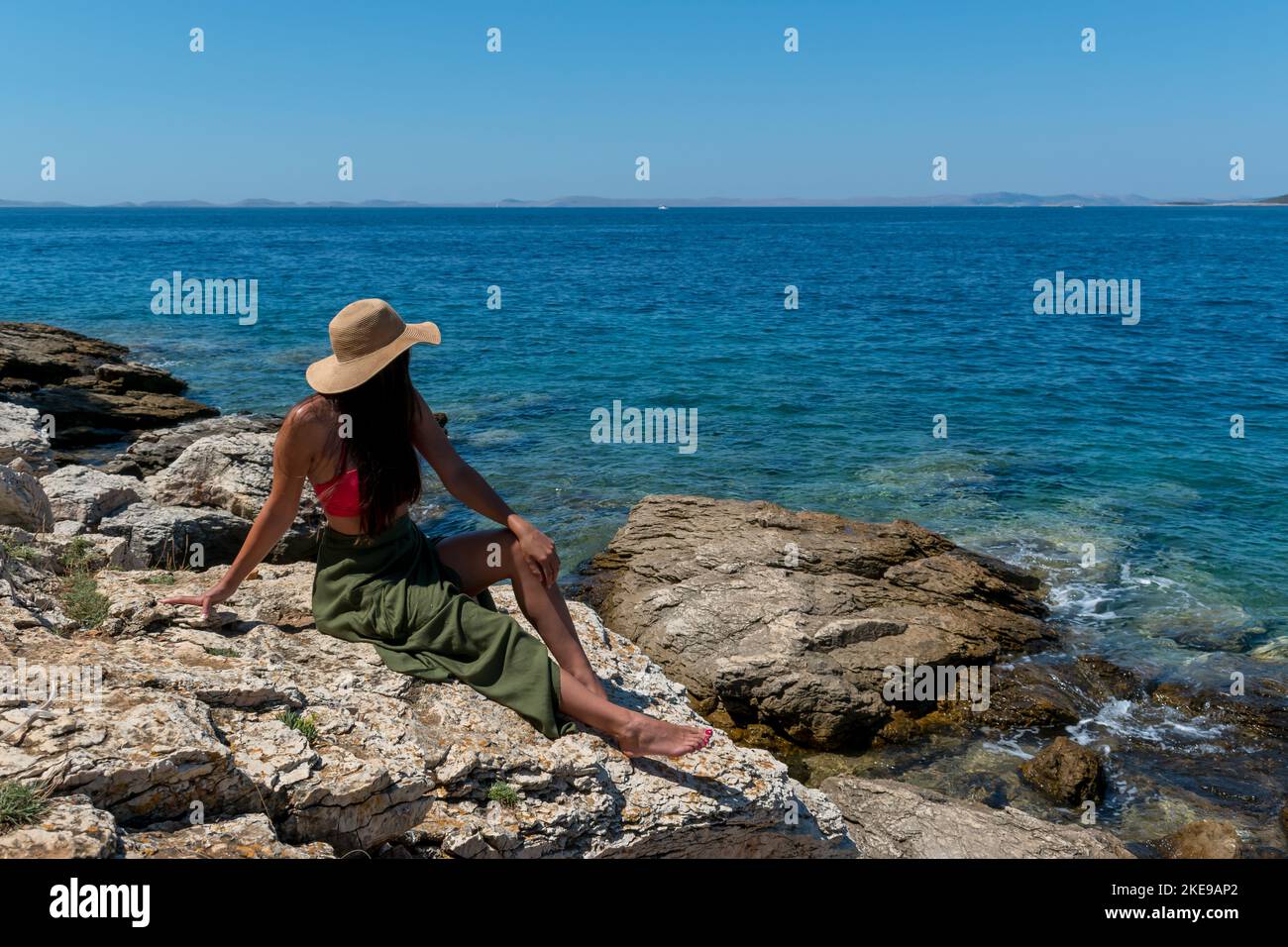 A back view of female wearing summer cloth sitting on rock in front of ...