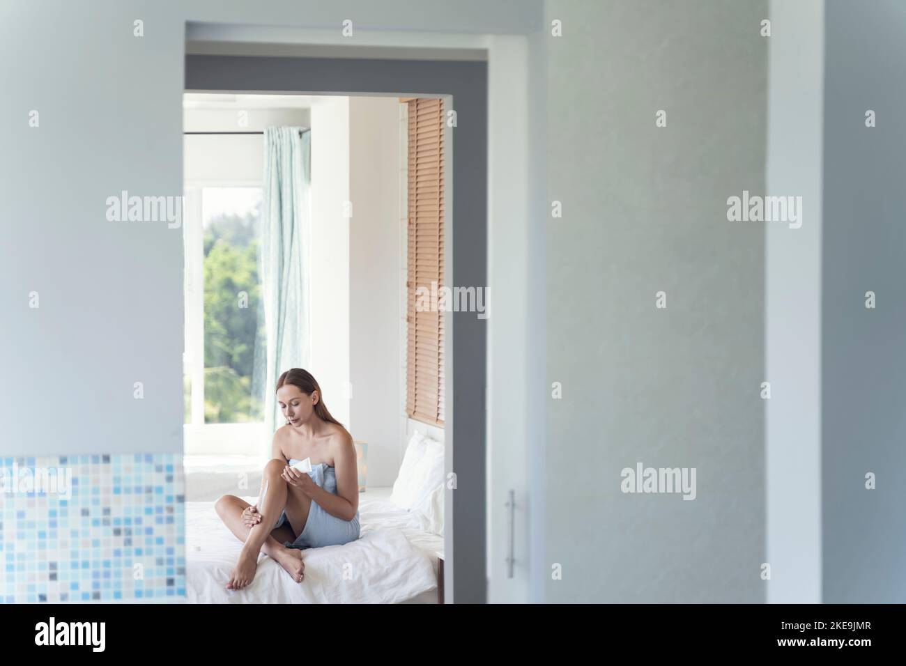 Young woman applies cream to her legs while sitting on the bed. A young woman makes body care while sitting on a bed in a bright room. Self care. Stock Photo