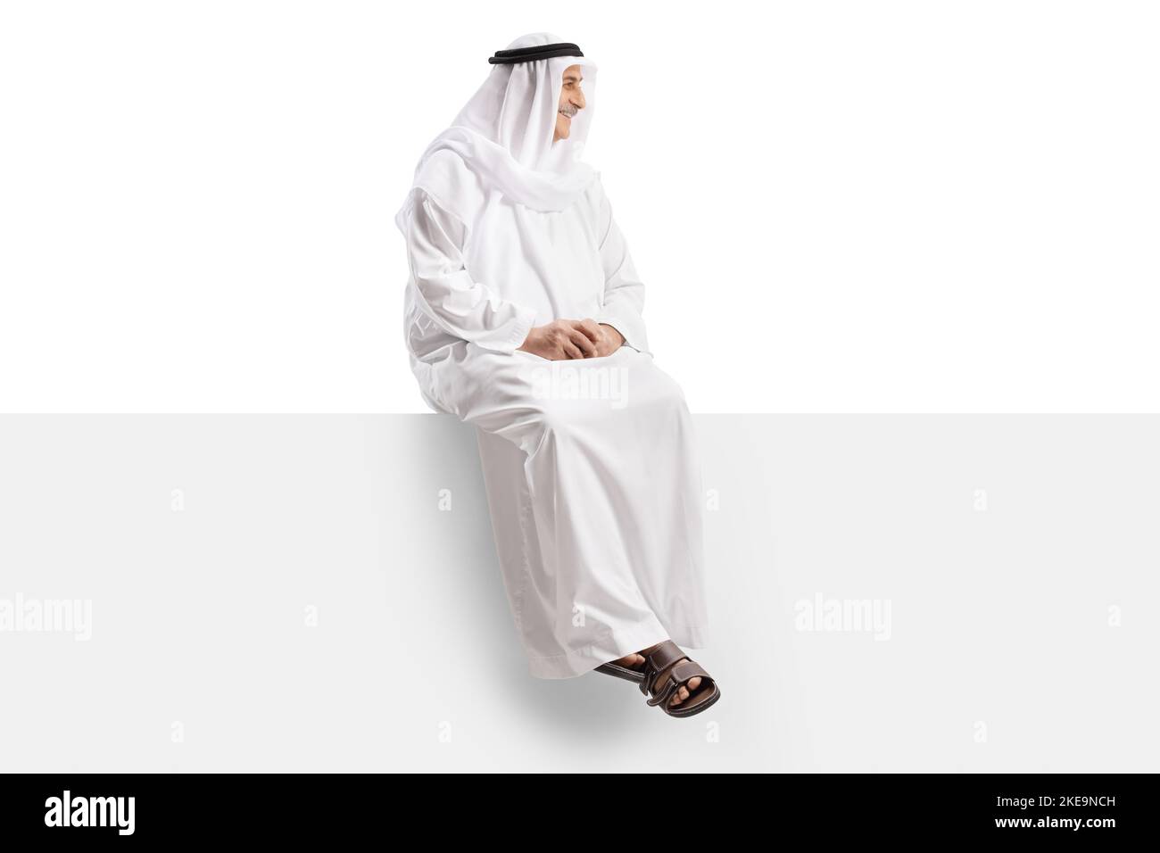 Arab man in an ethnic robe sitting on a blank panel and looking to the side isolated on white background Stock Photo