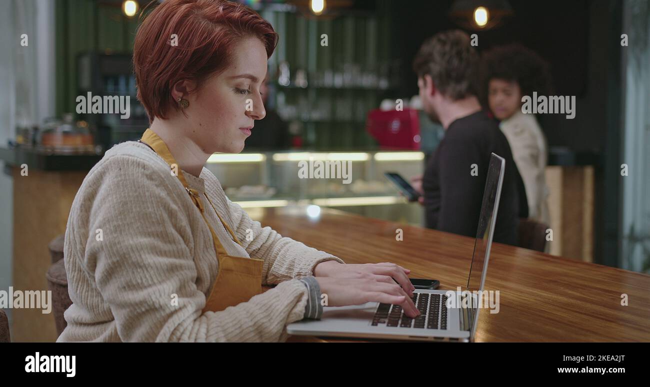 Young woman entrepreneur typing on laptop keyboard wearing apron seated ...