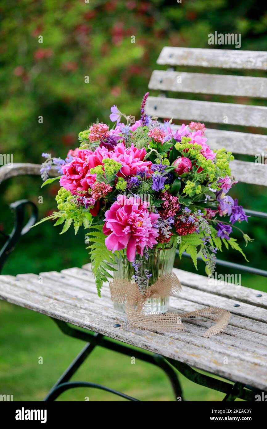 Bunter Blumenstrauss in rot, pink und violett Farbtönen mit Pfingstrosen und Akeleien, steht in Glas-Vase auf dekorativen Holz-Gartenbank Stock Photo