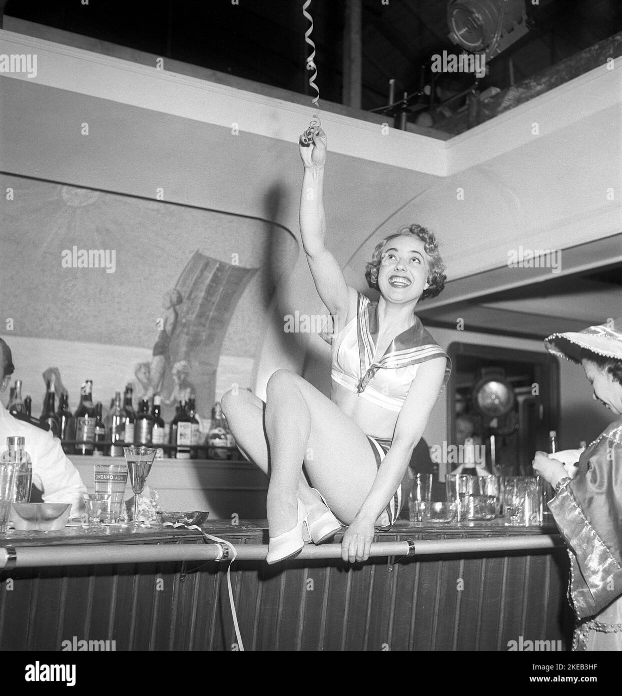 Party in the 1950s. A smiling young woman dressed in a costume however small, that resembles a sailor uniform, is sitting on top of the bar during a party at a filmstudio. Sweden 1950 Kristoffersson ref AX56-1 Stock Photo