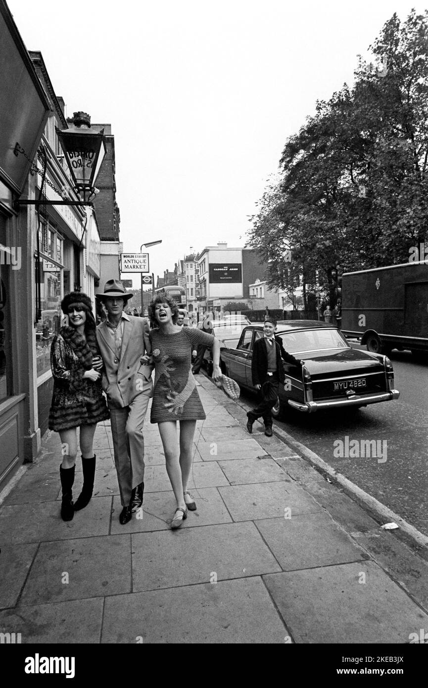 London in the 1960s. Ulla Larsson and Birgitta Bjerke at 251 Kings Road in front of the Antique market stalls. They are wearing the very typical 1960s fashion of their own design. The man's identity is unknown. Picture taken during the era of the Swinging Sixties, a youth-driven cultural revoluton in the UK in the mid to late 1960s. Note the typical hand patterned clothing.  London England October 1967. Stock Photo