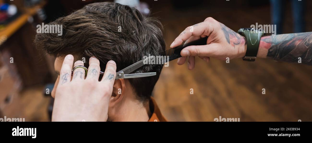 tattooed barber with comb and thinning scissors making hairstyle to brunette man, banner,stock image Stock Photo