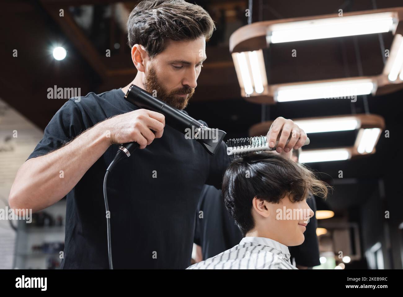Barber drying hair of teenage boy in hairdressing cape in beauty salon