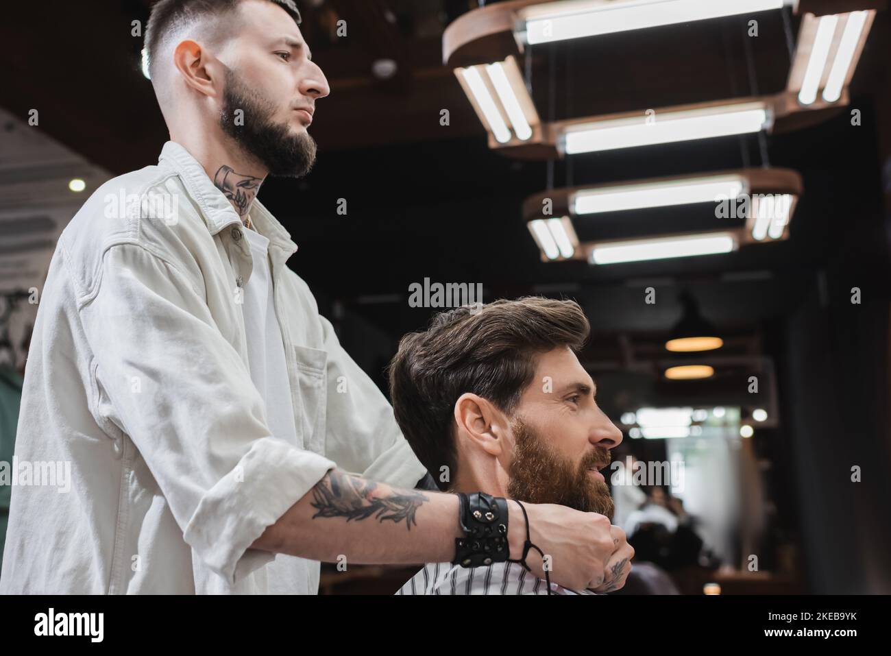 Side view of tattooed barber styling beard of smiling client in barbershop,stock image Stock Photo