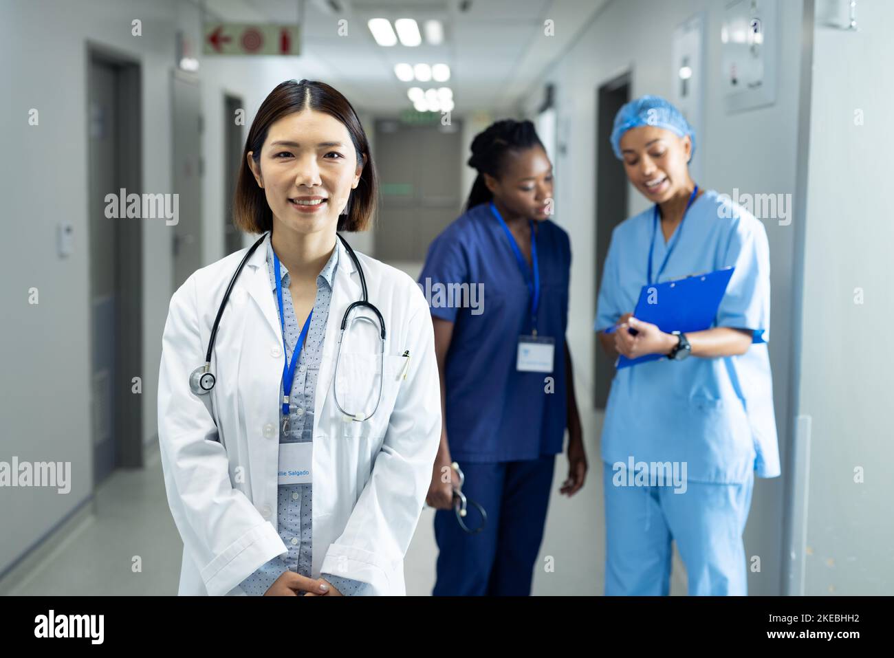 Portrait of smiling asian female doctor in hospital corridor, colleagues in background. Hospital, medical and healthcare services. Stock Photo
