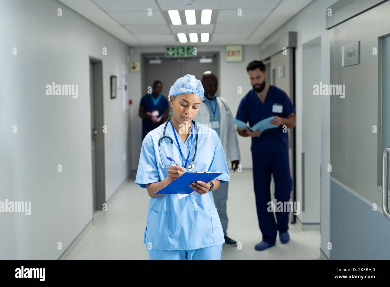 Biracial female doctor writing notes in busy hospital corridor, copy space. Hospital, medical and healthcare services. Stock Photo