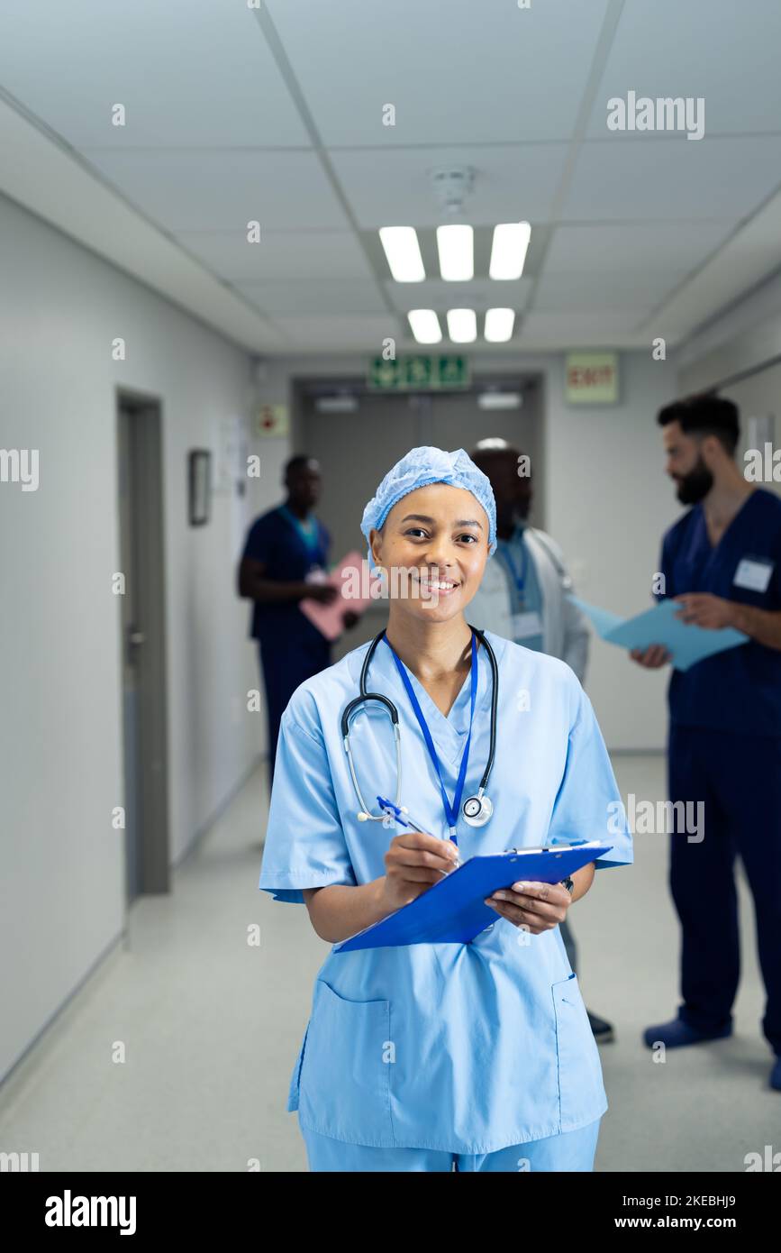 Vertical of smiling biracial female doctor writing notes in busy hospital corridor, copy space Stock Photo
