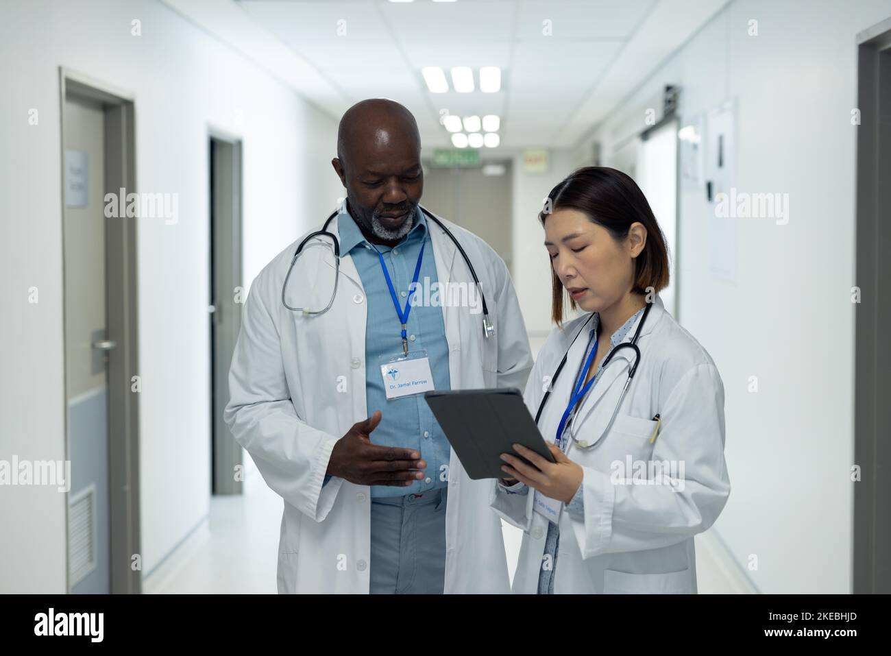 Two diverse female and female doctors looking at tablet and talking in hospital corridor. Hospital, medical and healthcare services. Stock Photo
