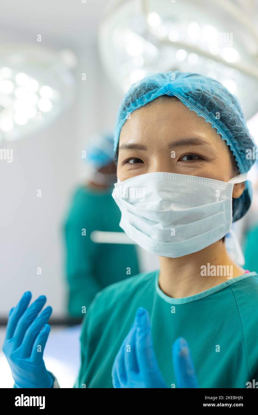 Vertical of smiling asian female surgeon in gown, mask, gloves and cap in theatre, copy space. Hospital, medical and healthcare services. Stock Photo