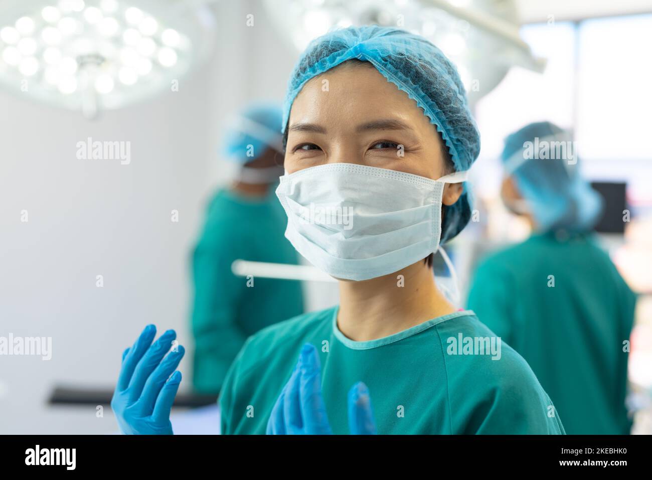 Smiling asian female surgeon in gown, mask, gloves and cap in operating theatre, copy space. Hospital, medical and healthcare services. Stock Photo
