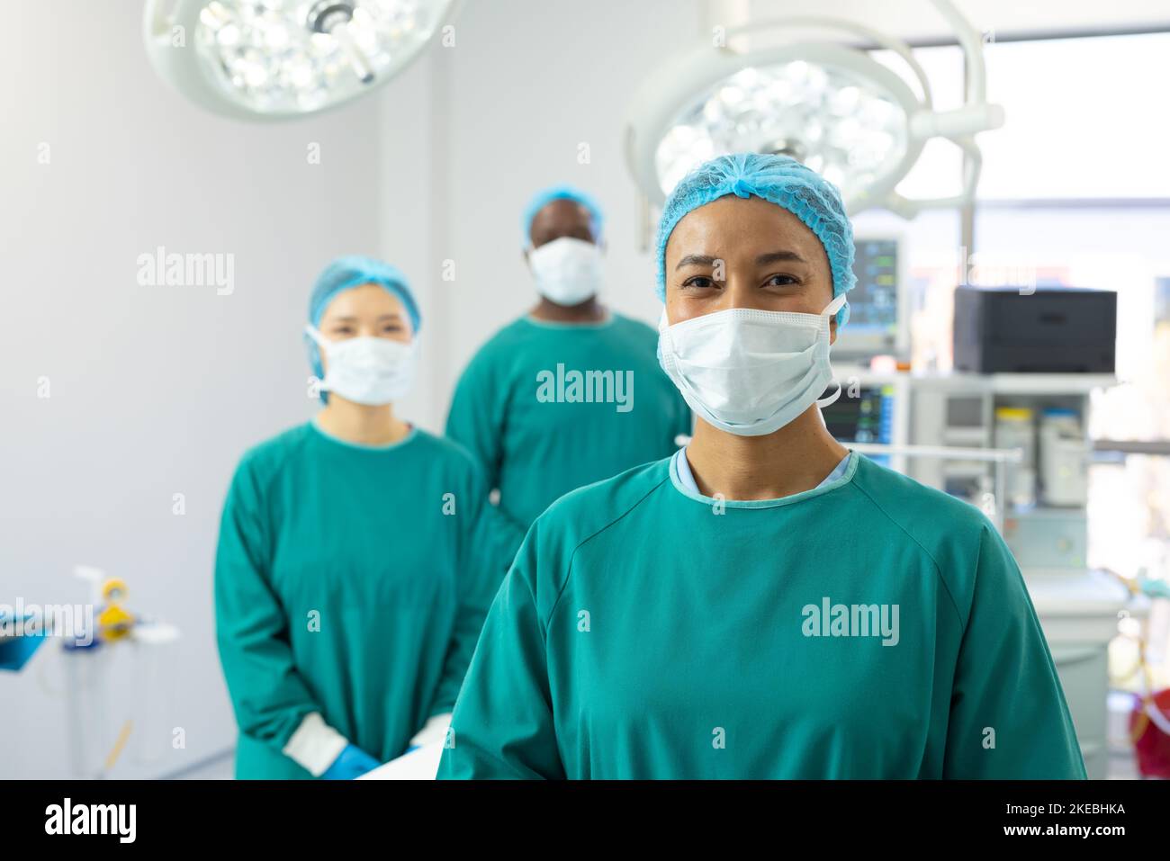 Portrait of smiling biracial female surgeon in mask and cap in operating theatre with colleagues. Hospital, medical and healthcare services. Stock Photo