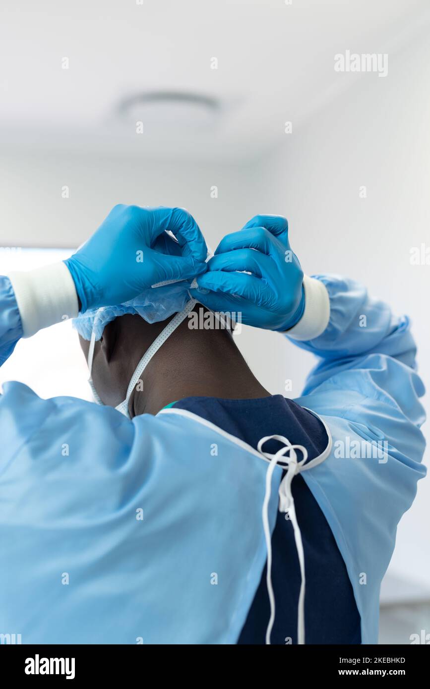 Rear of african american surgeon in gown and gloves tying on mask in theatre, vertical, copy space. Hospital, medical and healthcare services. Stock Photo