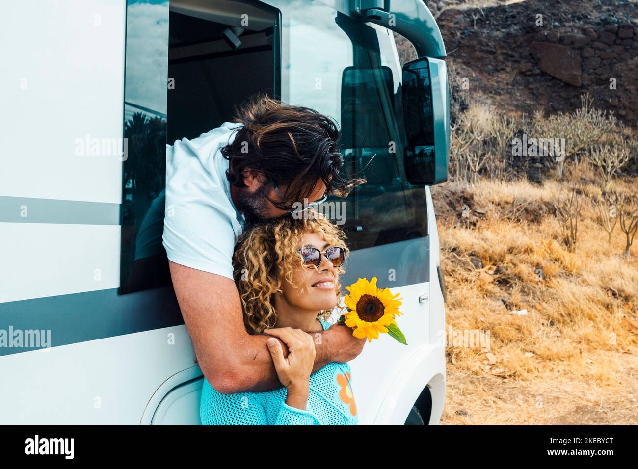 Man kissing woman and both enjoy travel lifestyle. Happy couple hug and smile in relationship against a modern camper van motor home. Alternative vacation on the road and freedom off grid life Stock Photo