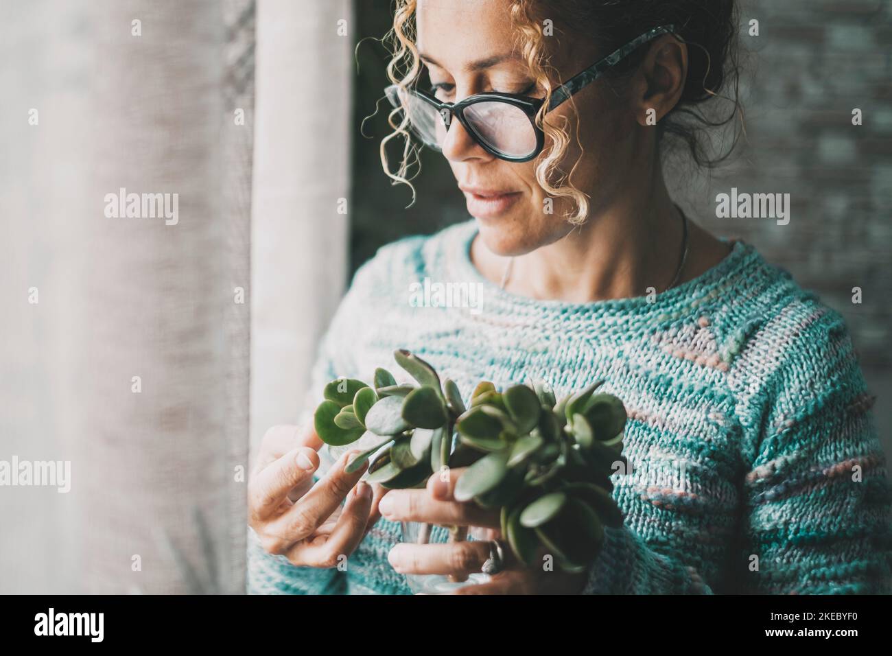 Close up portrait of young mature woman having care of a green plant at home in indoor leisure serene activity alone. Single lady enjoying life and apartment. One female people wearing eyewear indoor Stock Photo