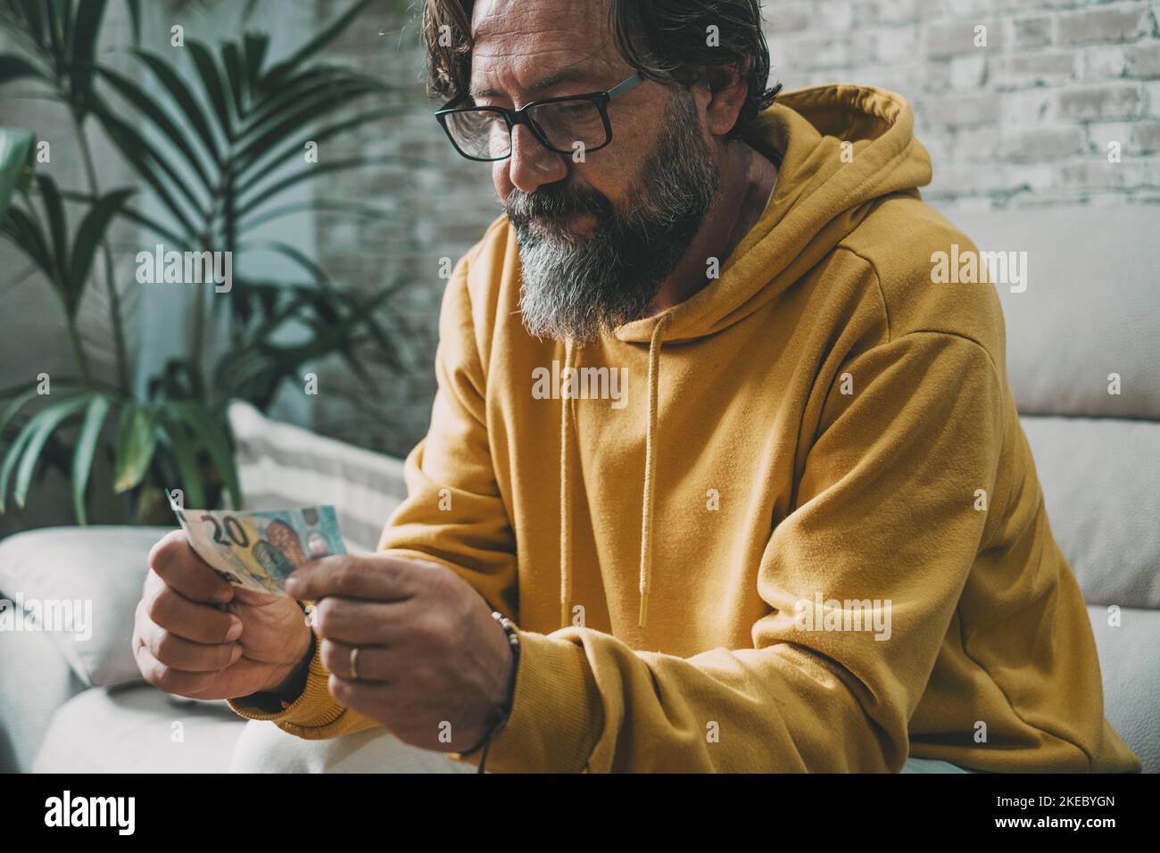Economy and money people problem concept. Man holding cash banknote with worried expression on face. Mature caucasian man looking last 20 euros sitting on the sofa. Real life Cash and bill payments Stock Photo